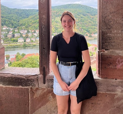 Madelyn McCullough stands by a window frame of an old stone building overlooking a river and lush mountains. 