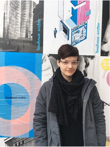 Samantha Shipeck wears a black and grey outfit and is standing alongside a wall with colorful posters. Caption: At the Bauhaus Archiv in Berlin during my time living in Rostock. Photo by Mary Lawrence.
