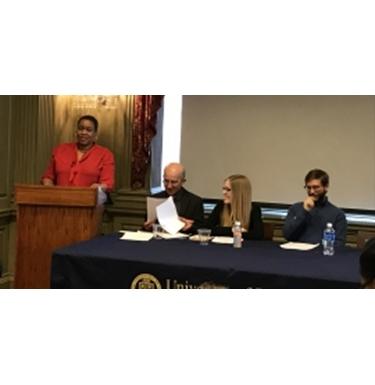 A speaker stands at a podium while a panel of three people sit at a table next to the podium.